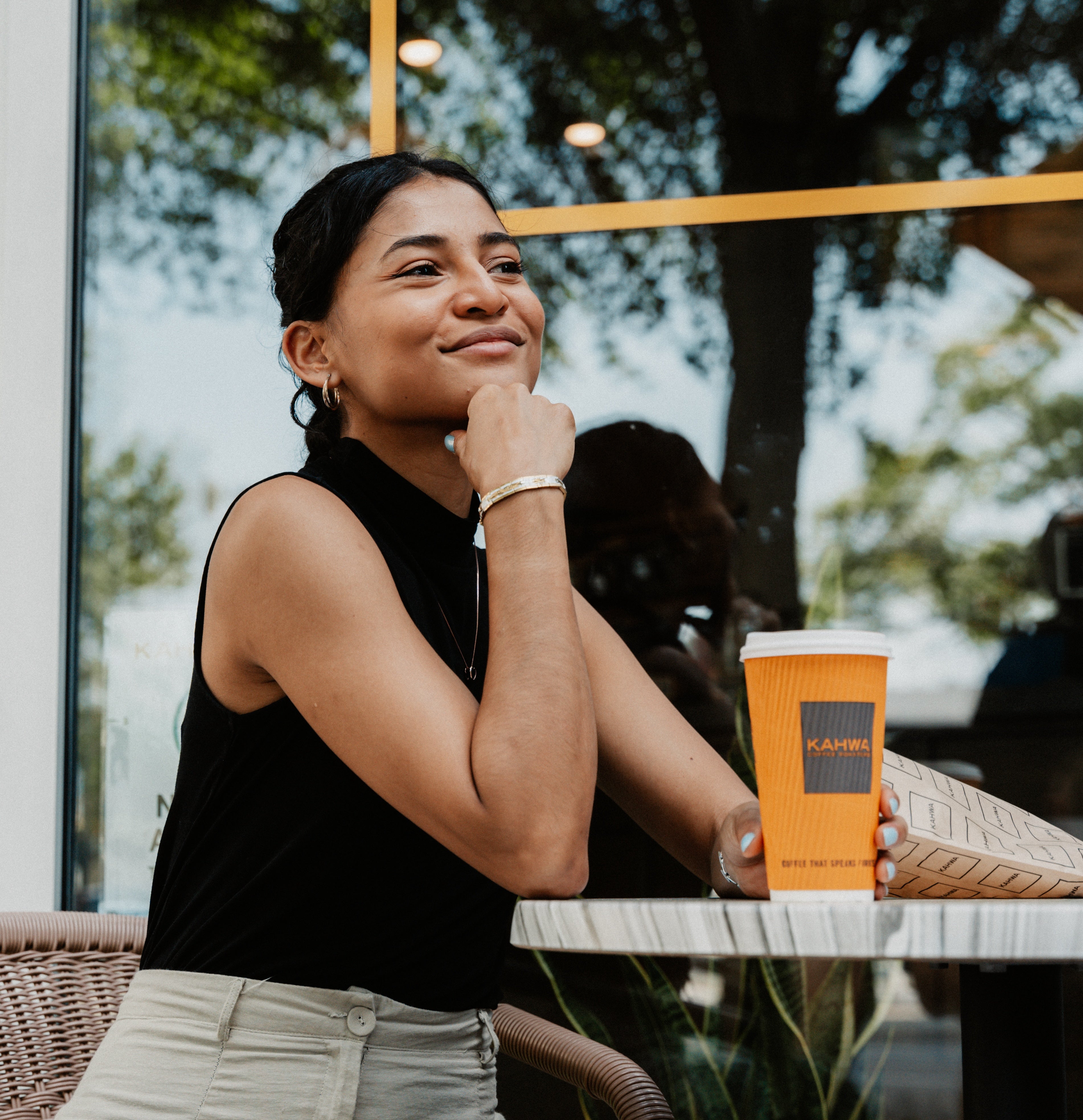 A guest smiling at an outdoor cafe table outside a Kahwa Coffee shop, holding a Kahwa to-go cup